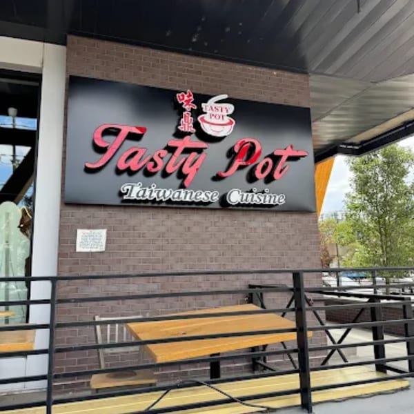 The exterior sign for Tasty Pot Taiwanese Cuisine, featuring red lettering on a black background, above outdoor seating.